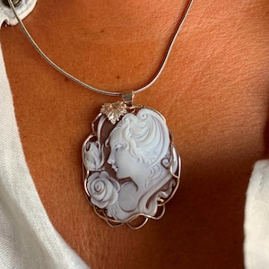 May include: A silver chain necklace with a cameo pendant featuring a woman's profile with a rose and leaves. The cameo is made of white shell and is set in a silver frame.
