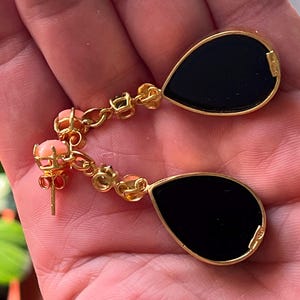 May include: Gold drop earrings with black, teardrop-shaped insets. The earrings have a chain of small coral-coloured beads and gold accents. The earrings are held in a hand.