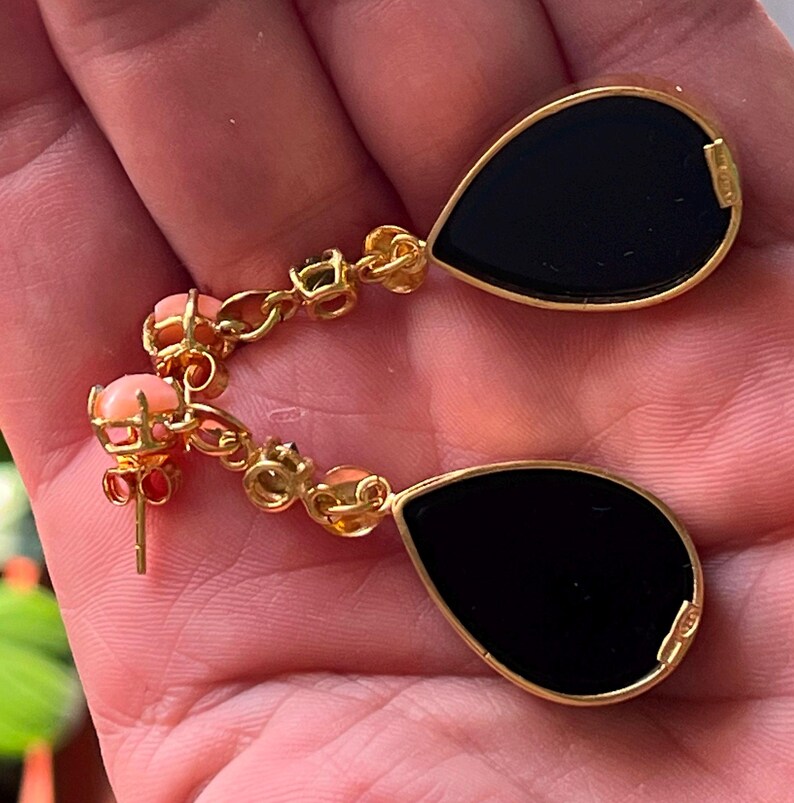 May include: Gold drop earrings with black, teardrop-shaped insets. The earrings have a chain of small coral-coloured beads and gold accents. The earrings are held in a hand.