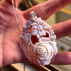 Op de afbeelding: Een bloemenhangerketting met een gebeeldhouwde, gebroken witte hanger met een roos en andere bloemmotieven. De hanger hangt aan een fijne zilveren ketting. De hanger is ongeveer 5 cm lang.