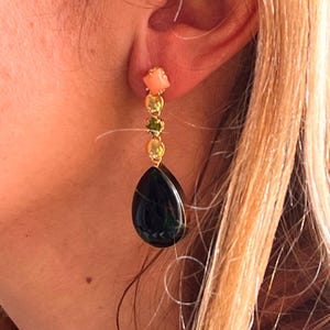 May include: A gold earring featuring a coral square, two gold circles, a green gem, and a black teardrop. The earring is displayed on an ear. The design is elegant and eye-catching.