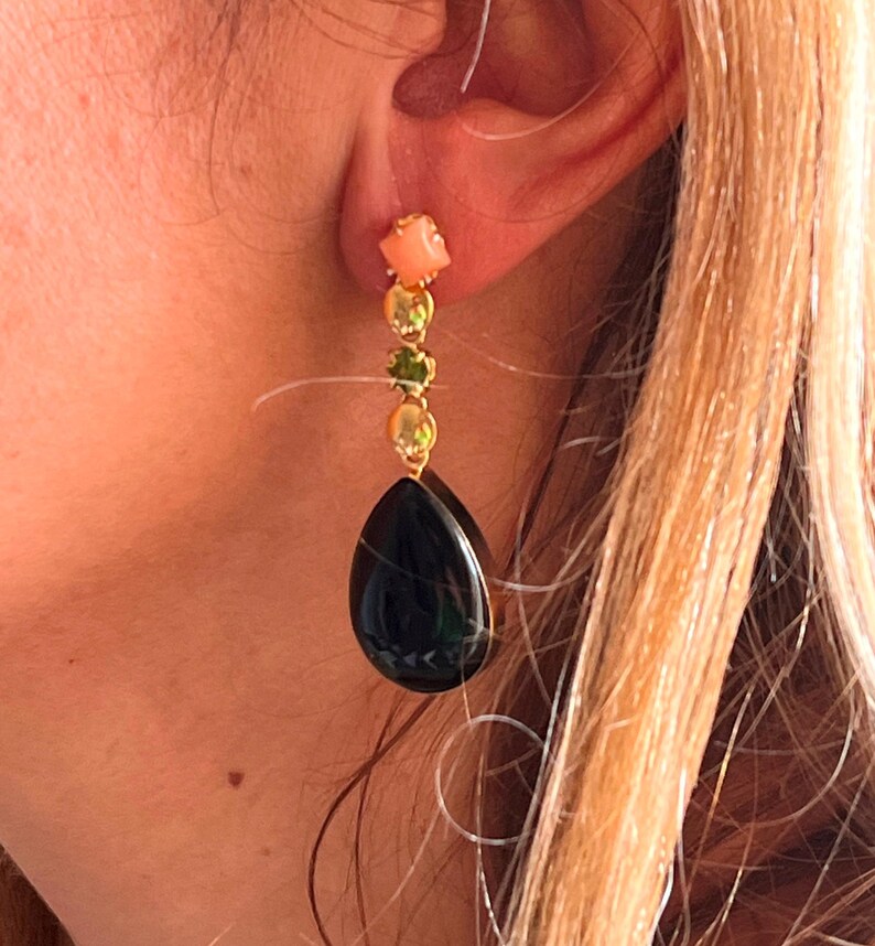May include: A gold earring featuring a coral square, two gold circles, a green gem, and a black teardrop. The earring is displayed on an ear. The design is elegant and eye-catching.