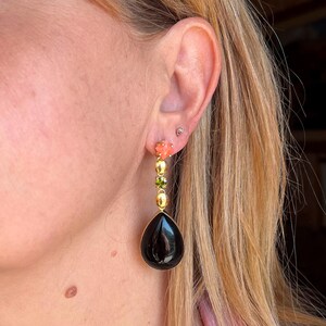 May include: Gold drop earrings with a black, teardrop-shaped stone. The earrings have coral-coloured flower accents, green and gold beads. The earrings are worn on a person's ear.