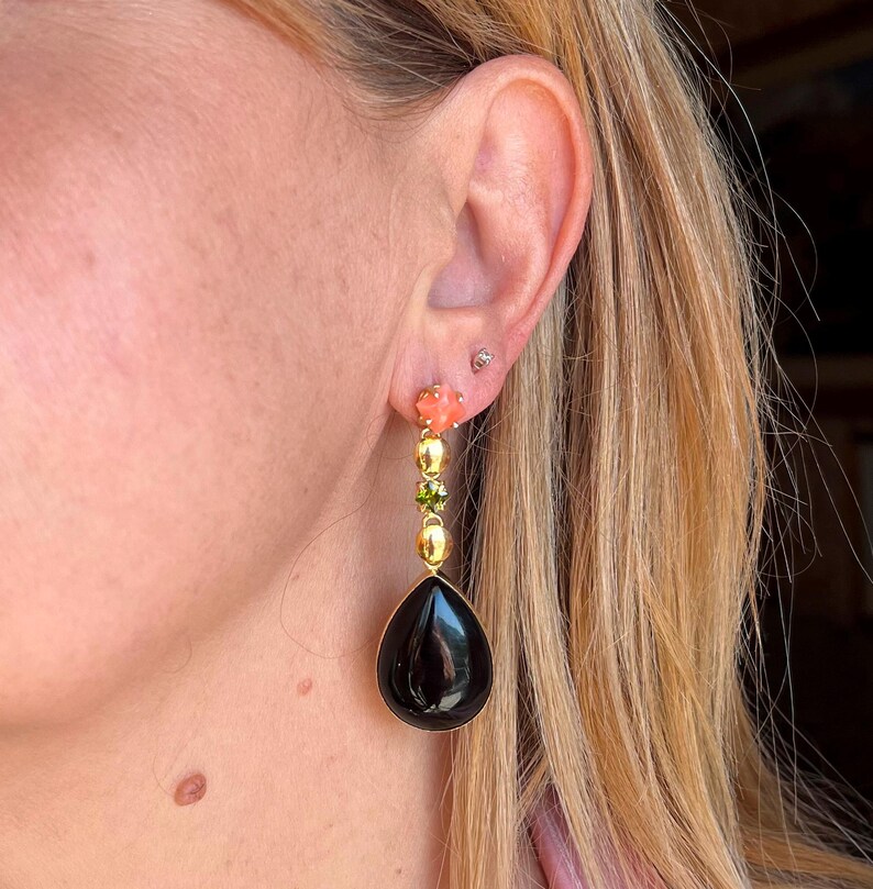 May include: Gold drop earrings with a black, teardrop-shaped stone. The earrings have coral-coloured flower accents, green and gold beads. The earrings are worn on a person's ear.