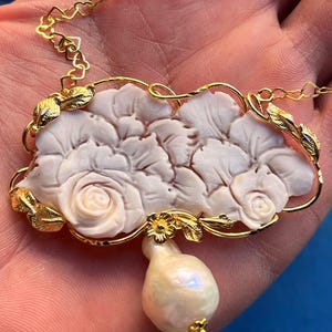 Puede incluir: Un collar dorado con un colgante floral blanco tallado, con detalles de rosas y hojas. Una gran perla en forma de pera cuelga debajo del colgante. La cadena tiene eslabones en forma de coraz&oacute;n.