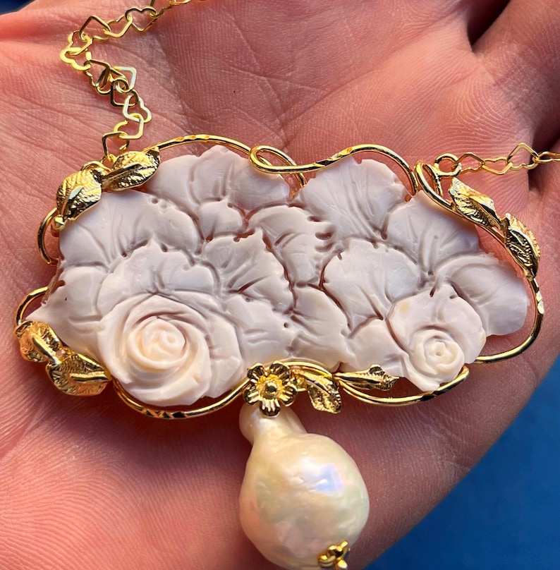 Puede incluir: Un collar dorado con un colgante floral blanco tallado, con detalles de rosas y hojas. Una gran perla en forma de pera cuelga debajo del colgante. La cadena tiene eslabones en forma de coraz&oacute;n.