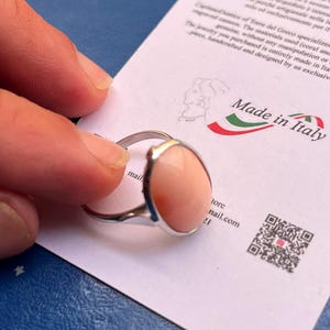 K&ouml;nnte beinhalten: Ein silberner Ring mit einem gro&szlig;en, ovalen, pfirsichfarbenen Stein. Der Ring wird &uuml;ber einer wei&szlig;en Karte mit der Aufschrift "Made in Italy" und anderem Text gehalten. Der Ring ist handgefertigt und in Italien entworfen.