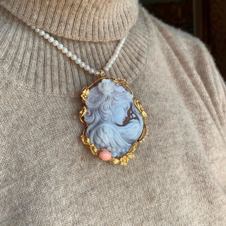 May include: A white cameo pendant with a gold frame and floral details. The cameo features a woman's profile with a delicate floral design. The pendant is hanging from a pearl necklace.