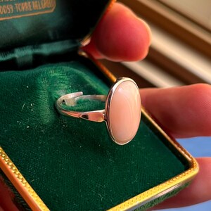 K&ouml;nnte beinhalten: Ein silberner Ring mit einem ovalen, hellrosa Edelstein. Der Ring ist in einem silbernen Band gefasst und wird in einer gr&uuml;nen Samt-Schmuckschatulle pr&auml;sentiert. Der Edelstein hat eine glatte, polierte Oberfl&auml;che.