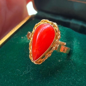 Puede incluir: Un anillo de oro con una piedra de coral roja brillante en forma de lágrima. El coral está engastado en un marco de oro con un diseño de cuerda trenzada. El anillo se muestra sobre una superficie de terciopelo verde.