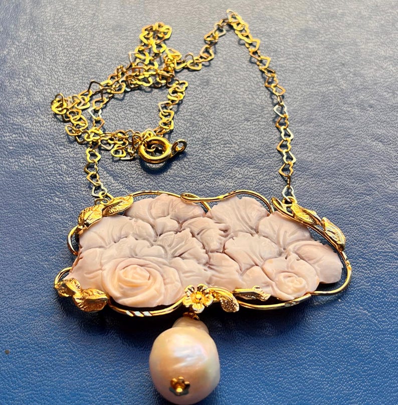 Puede incluir: Un collar dorado con un colgante de rosas rosas talladas y una perla colgante. El colgante est&aacute; enmarcado por un borde dorado con detalles de hojas. El collar tiene una cadena y un cierre delicados.