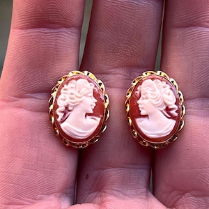May include: A pair of gold tone cameo earrings with a red cameo featuring a white profile of a woman.