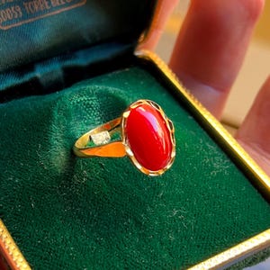 Op de afbeelding: Een gouden ring met een grote, ovale, helderrode steen. De ring is gezet in een gouden band met aan weerszijden van de rode steen een kleine, vierkante, heldere steen. De ring wordt gepresenteerd in een groene fluwelen sieradendoos.