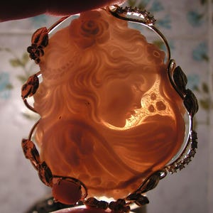 May include: A cameo pendant featuring a carved image of a woman's face, set in a gold-tone metal frame with a floral design and clear rhinestones.