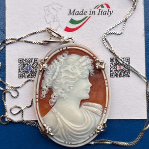 May include: Oval cameo pendant necklace featuring a white profile carving set against a brown background, framed with a white border and small diamonds. The pendant hangs from a silver chain. The words "Made in Italy" are visible.