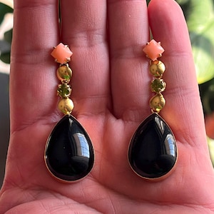 May include: A pair of gold-toned drop earrings. Each earring features a peach-coloured square-shaped stone at the top, followed by gold beads and green stones. The bottom of each earring has a large, black, teardrop-shaped stone.
