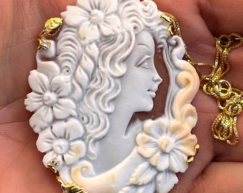 May include: Oval pendant necklace featuring a white cameo of a woman's profile, adorned with floral and leaf accents. The pendant is suspended from a delicate gold chain. The cameo is approximately 2 inches in length.