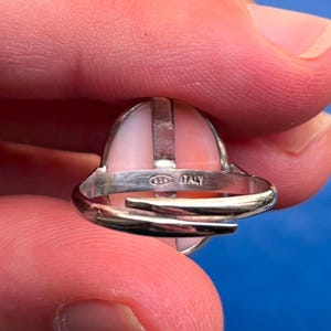 K&ouml;nnte beinhalten: Ein Silberring mit einem hellrosa, ovalen Stein. Der Ring hat ein geteiltes Schaftdesign und ist mit "925 ITALY" gestempelt. Der Stein ist in einer L&uuml;nettenfassung gefasst.
