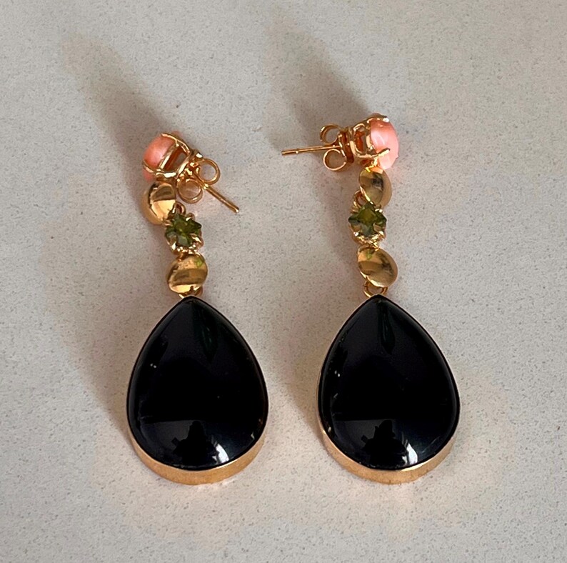 May include: A pair of gold drop earrings. Each earring has a black, teardrop-shaped stone with a gold rim, and a series of smaller green and gold stones, topped with a pink stone. The earrings are for pierced ears.
