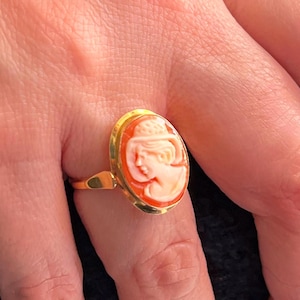 May include: A gold ring featuring an oval cameo with a profile of a woman. The cameo is peach-colored, set in a gold bezel, and mounted on a gold band. The ring is a classic piece of jewelry.