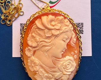 Puede incluir: Un collar dorado con un colgante de camafeo ovalado. El camafeo presenta un perfil tallado de una mujer con flores, engastado en un marco dorado. El colgante está sujeto a una delicada cadena dorada. La etiqueta dice "Made in Italy".