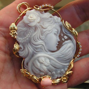 May include: A gold pendant featuring a carved shell cameo of a woman's profile with flowing hair and a rose. The cameo is surrounded by a gold frame with leaves and flowers, and a pink stone is at the bottom.