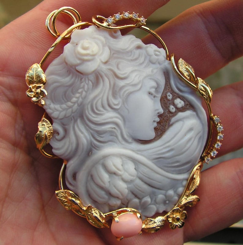 May include: A gold pendant featuring a carved shell cameo of a woman's profile with flowing hair and a rose. The cameo is surrounded by a gold frame with leaves and flowers, and a pink stone is at the bottom.