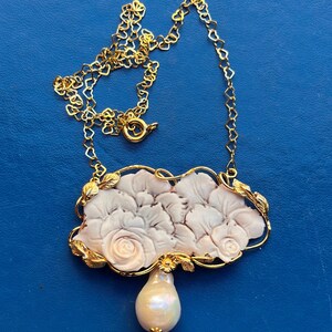 Puede incluir: Un collar dorado con un colgante con un dise&ntilde;o floral tallado y una gota de perla en forma de pera. La cadena del collar tiene eslabones en forma de coraz&oacute;n. El colgante tiene un marco dorado con detalles de hojas.