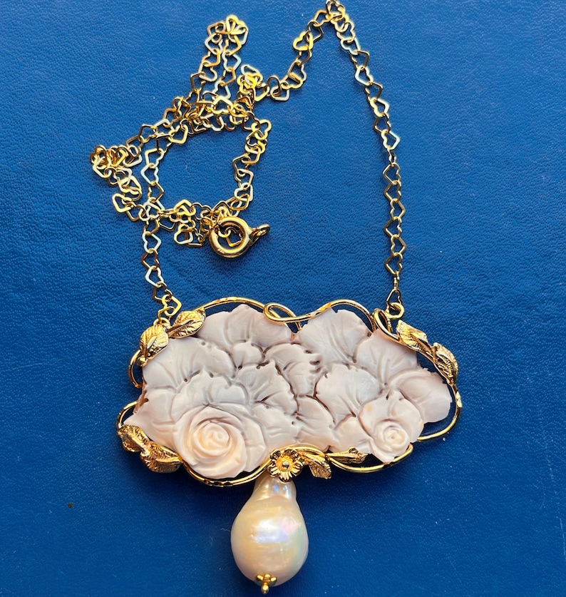 Puede incluir: Un collar dorado con un colgante con un dise&ntilde;o floral tallado y una gota de perla en forma de pera. La cadena del collar tiene eslabones en forma de coraz&oacute;n. El colgante tiene un marco dorado con detalles de hojas.