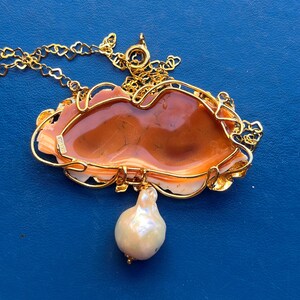 Puede incluir: Un collar dorado con un colgante ornamentado. El colgante presenta una piedra central de color marr&oacute;n anaranjado enmarcada por un dise&ntilde;o de filigrana dorada. Una gran perla en forma de pera cuelga de la parte inferior. El collar est&aacute; sobre un fondo azul.