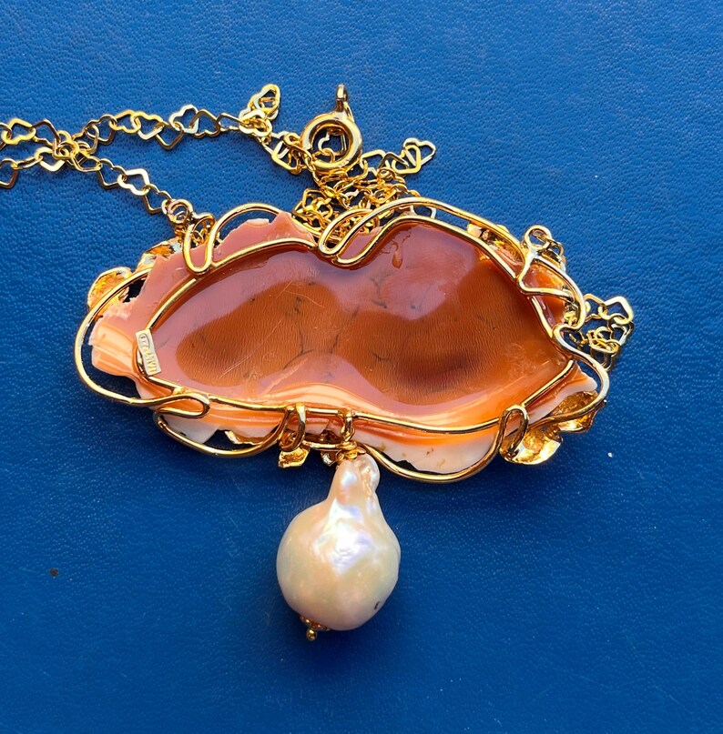 Puede incluir: Un collar dorado con un colgante ornamentado. El colgante presenta una piedra central de color marr&oacute;n anaranjado enmarcada por un dise&ntilde;o de filigrana dorada. Una gran perla en forma de pera cuelga de la parte inferior. El collar est&aacute; sobre un fondo azul.
