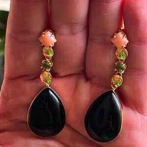 May include: Gold drop earrings with a teardrop-shaped black stone, complemented by peach and green gemstones. The earrings have a delicate design with a gold chain.