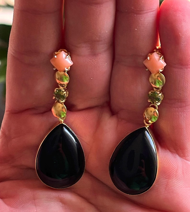 May include: Gold drop earrings with a teardrop-shaped black stone, complemented by peach and green gemstones. The earrings have a delicate design with a gold chain.