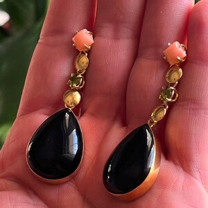 May include: A pair of gold-toned drop earrings. Each earring features a black teardrop-shaped stone, accented with coral, green, and gold-toned beads. The earrings are held in a hand.