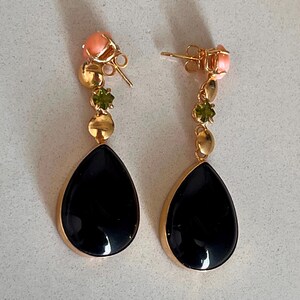 May include: A pair of gold-coloured drop earrings. Each earring features a black teardrop-shaped stone, accented with gold, peach, and green gemstones. The earrings have a post-back closure.