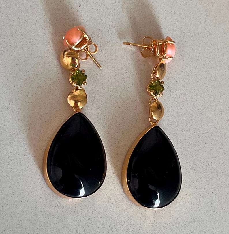 May include: A pair of gold-coloured drop earrings. Each earring features a black teardrop-shaped stone, accented with gold, peach, and green gemstones. The earrings have a post-back closure.
