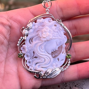 Puede incluir: Un colgante de plata con un camafeo de concha tallada que representa el rostro de una mujer con detalles florales. El camafeo está rodeado por un marco de plata con piedras preciosas verdes y hojas de plata.