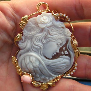 May include: A delicate cameo pendant featuring a carved woman's face with flowing hair, set in a gold frame with leaves and a pink coral accent.