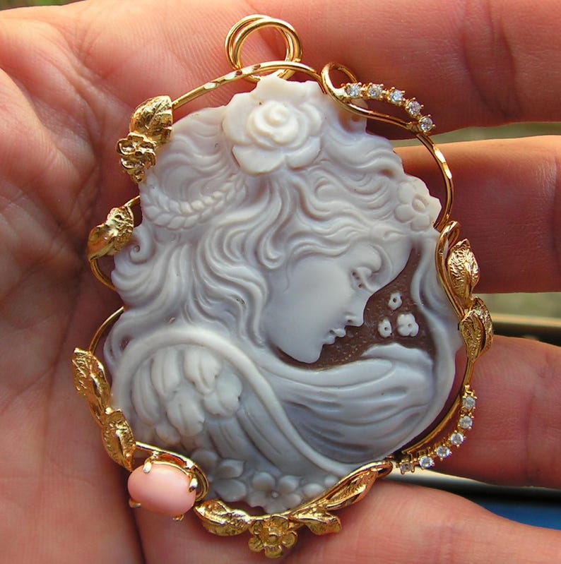May include: A delicate cameo pendant featuring a carved woman's face with flowing hair, set in a gold frame with leaves and a pink coral accent.