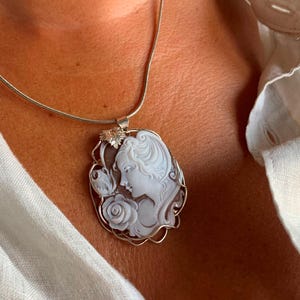 May include: A silver chain necklace with a cameo pendant featuring a woman's profile with a rose and a butterfly. The cameo is set in a silver frame.