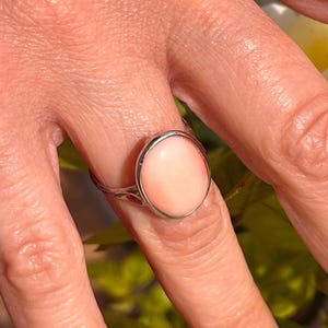 K&ouml;nnte beinhalten: Ein Silberring mit einem gro&szlig;en, ovalen, hellrosa Edelstein. Der Edelstein ist in einem polierten Silberband gefasst. Der Ring wird an einem Finger gezeigt, mit einem verschwommenen Hintergrund aus gr&uuml;nen Bl&auml;ttern.