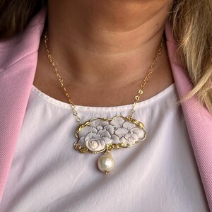 Puede incluir: Un collar dorado con un colgante en forma de nube adornado con dise&ntilde;os florales grises y blancos. Una gran perla en forma de pera cuelga debajo. El collar se lleva sobre una camiseta blanca y una blazer rosa.