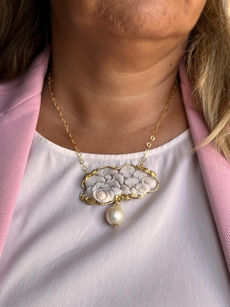 Puede incluir: Un collar dorado con un colgante en forma de nube adornado con dise&ntilde;os florales grises y blancos. Una gran perla en forma de pera cuelga debajo. El collar se lleva sobre una camiseta blanca y una blazer rosa.