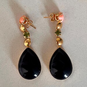May include: A pair of gold drop earrings. Each earring has a black, teardrop-shaped stone, gold beads, green stones, and a pink coral-like stud.
