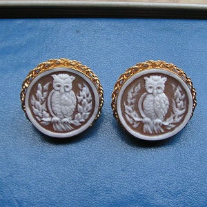 May include: A pair of gold tone earrings with a cameo of a white owl on a brown background. The owl is perched on a branch with leaves.