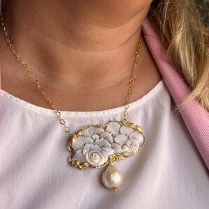 Puede incluir: Collar dorado con un colgante con dise&ntilde;o floral en blanco y gris. El colgante tiene una gran perla en forma de pera. La cadena tiene eslabones en forma de coraz&oacute;n. Una joya elegante.