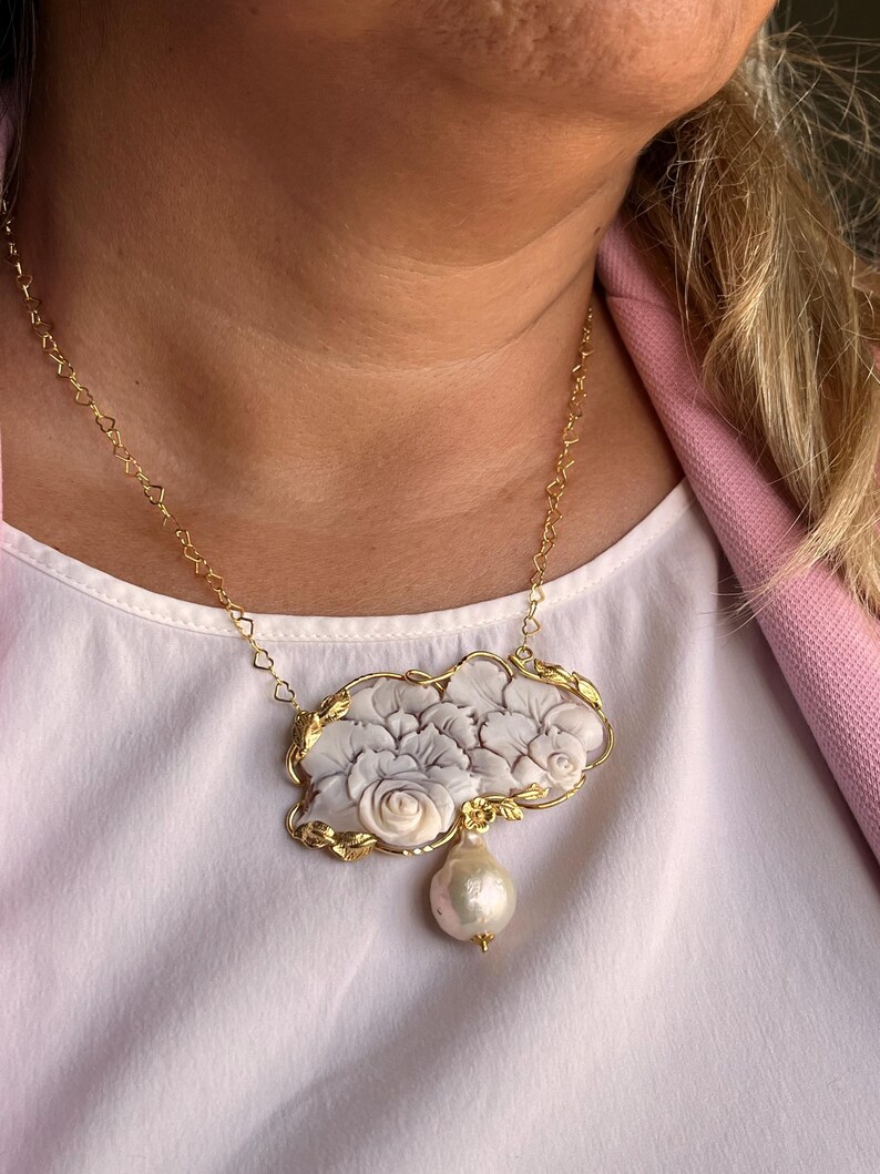 Puede incluir: Collar dorado con un colgante con dise&ntilde;o floral en blanco y gris. El colgante tiene una gran perla en forma de pera. La cadena tiene eslabones en forma de coraz&oacute;n. Una joya elegante.