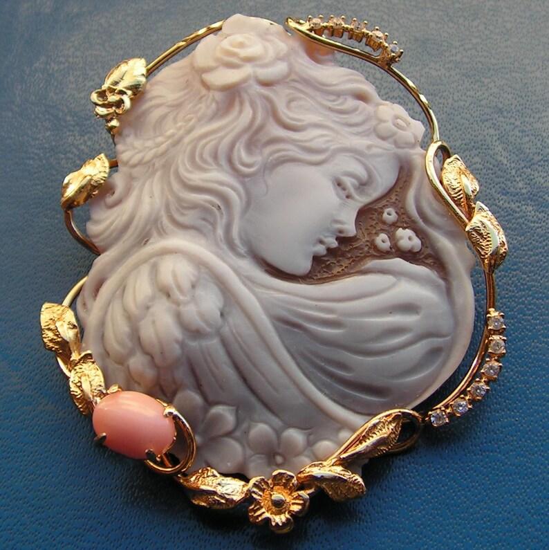 May include: A cameo brooch featuring a carved shell of a woman's profile with a rose in her hair. The cameo is set in a gold tone metal frame with a delicate floral design and clear rhinestones. A pink shell cameo is also included in the design.
