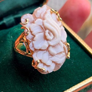 Puede incluir: Un anillo de oro con un diseño de flor rosa pálido tallada. La flor tiene pétalos detallados y está adornada con filigrana dorada. El anillo se encuentra sobre un fondo verde, resaltando los intrincados detalles del anillo.