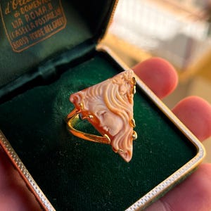 May include: A gold ring with a triangular, peach-colored cameo featuring a classical female profile. The ring is set in a gold band and displayed in a green velvet jewelry box.
